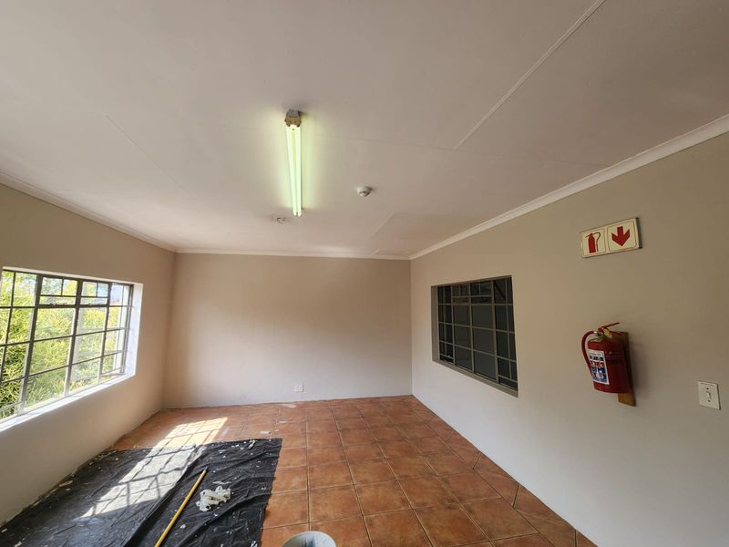A clean, empty office space featuring tiled flooring, steel-framed windows, and essential fire safety equipment, suitable for a professional workspace.