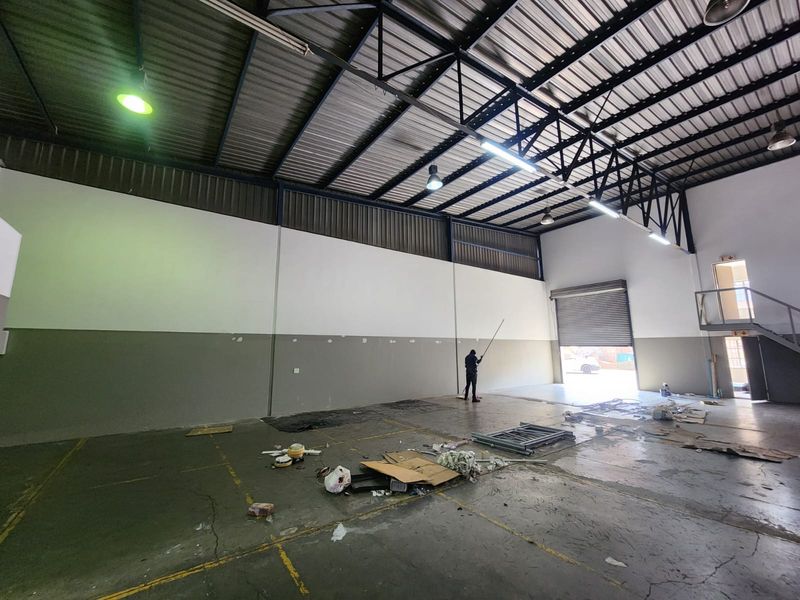 Spacious industrial warehouse unit featuring high ceilings with exposed steel trusses, concrete flooring, and a large roller shutter door for easy access.