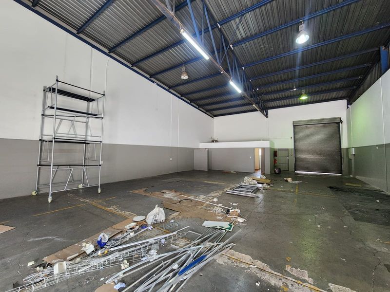 Spacious industrial warehouse unit featuring high ceilings with exposed steel trusses, concrete flooring, and a large roller shutter door for easy access.