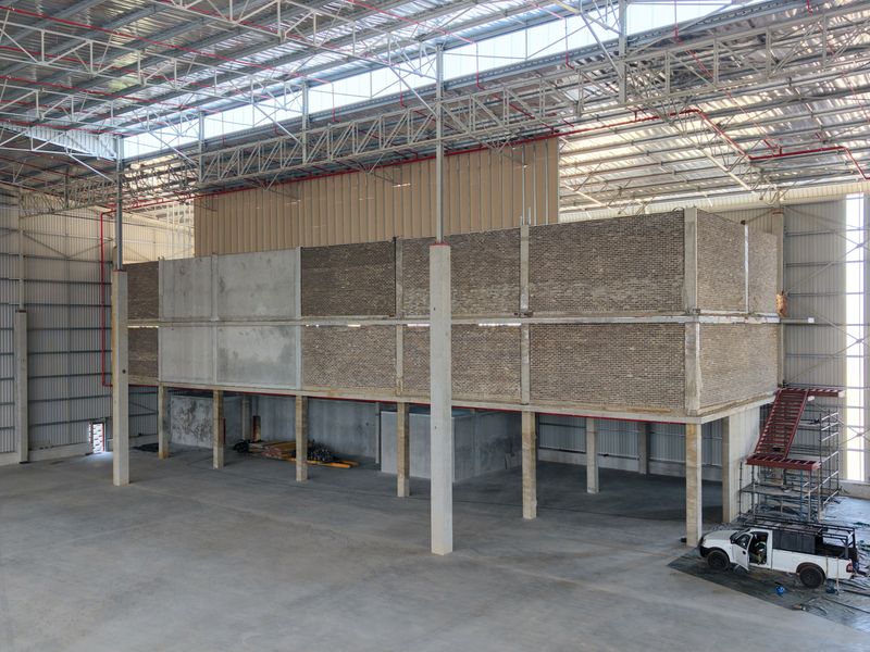Large industrial warehouse space featuring a high-clearance steel portal frame, concrete flooring, and an unfinished mezzanine level with brick and concrete structural elements.