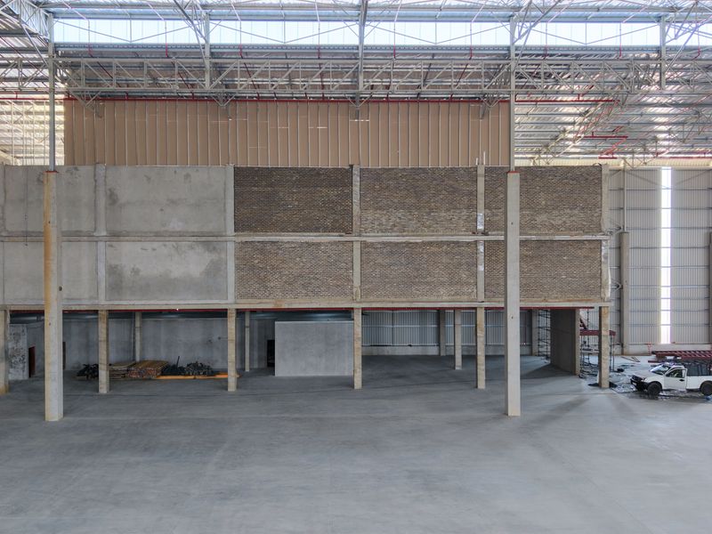 A large, open-plan industrial warehouse space featuring high ceilings with exposed steel trusses, concrete flooring, and structural brick and concrete wall elements.