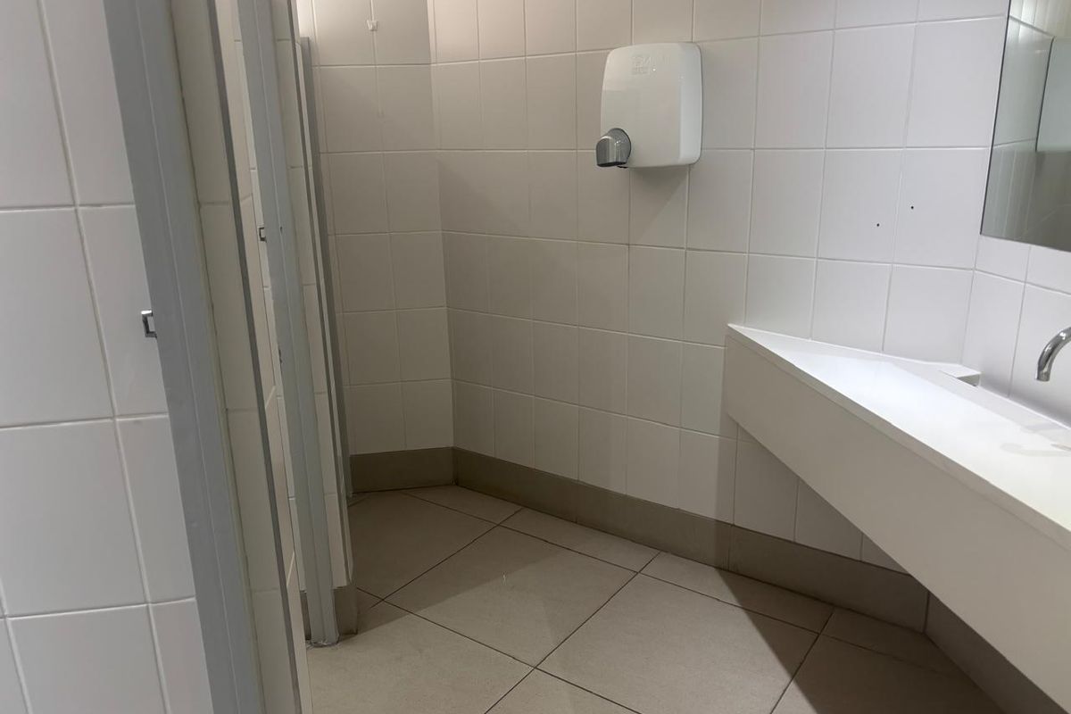 A functional office restroom featuring tiled walls and flooring, a wall-mounted hand dryer, and a built-in vanity basin with partitioned cubicles.