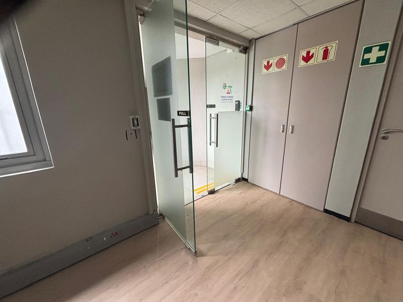 A clean, professional office entrance featuring a modern frosted glass door, light wood-look laminate flooring, and integrated storage cupboards with safety signage.