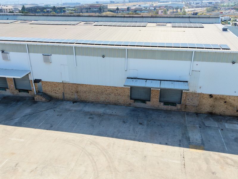 Aerial view of an industrial warehouse facility featuring a large metal roof with solar panels, multiple loading bays, and a spacious concrete yard.