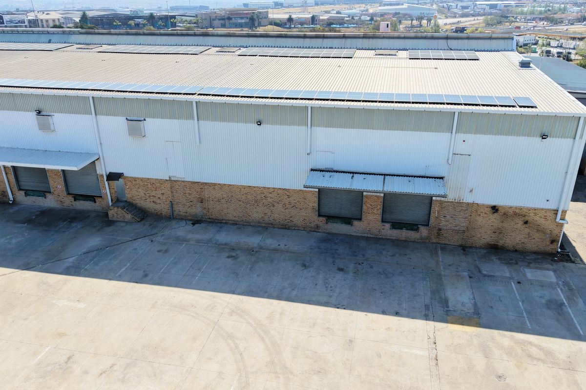 Aerial view of an industrial warehouse facility featuring a large metal roof with solar panels, multiple loading bays, and a spacious concrete yard.