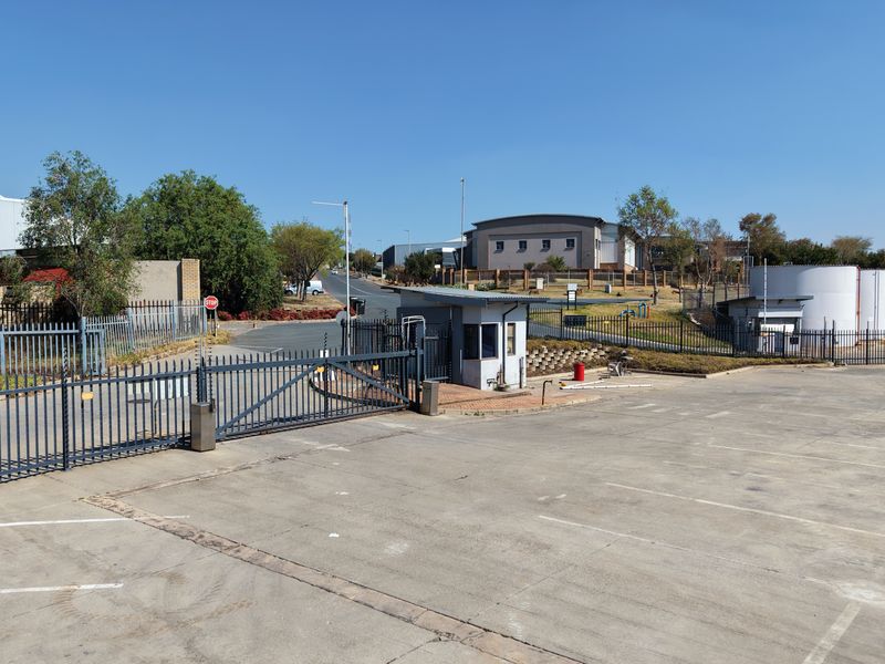 Industrial property entrance featuring a secure gated access point, guard house, and extensive paved parking area with perimeter security fencing.
