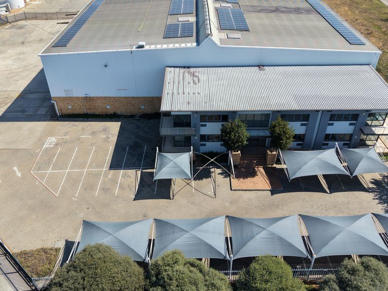 Aerial view of an industrial property featuring a large warehouse with solar panels, paved parking area, and multiple shade sail structures.