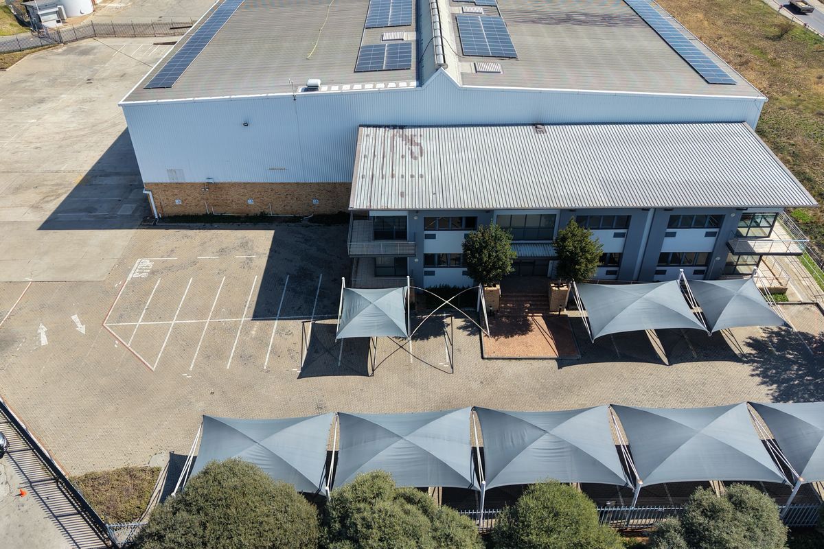 Aerial view of an industrial property featuring a large warehouse with solar panels, paved parking area, and multiple shade sail structures.