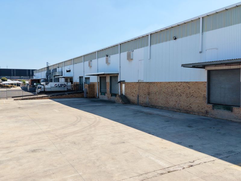 Industrial warehouse exterior featuring multiple loading docks with roller shutter doors, a large concrete yard, and secure perimeter fencing.