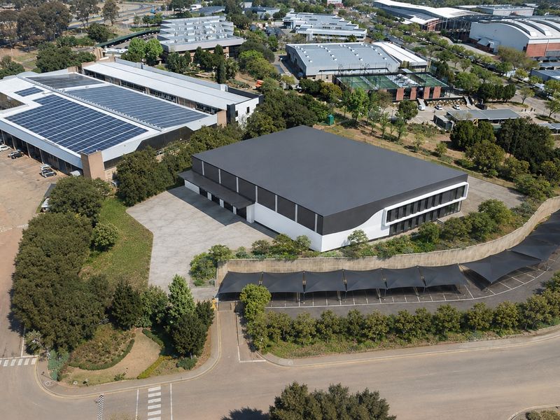 Aerial view of a modern industrial property featuring a large warehouse with solar panels, paved parking, and established landscaping in a business park setting.