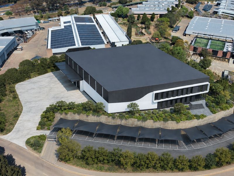 Aerial view of a modern industrial facility featuring a large warehouse with solar panels, extensive paved parking with shade structures, and landscaped grounds.