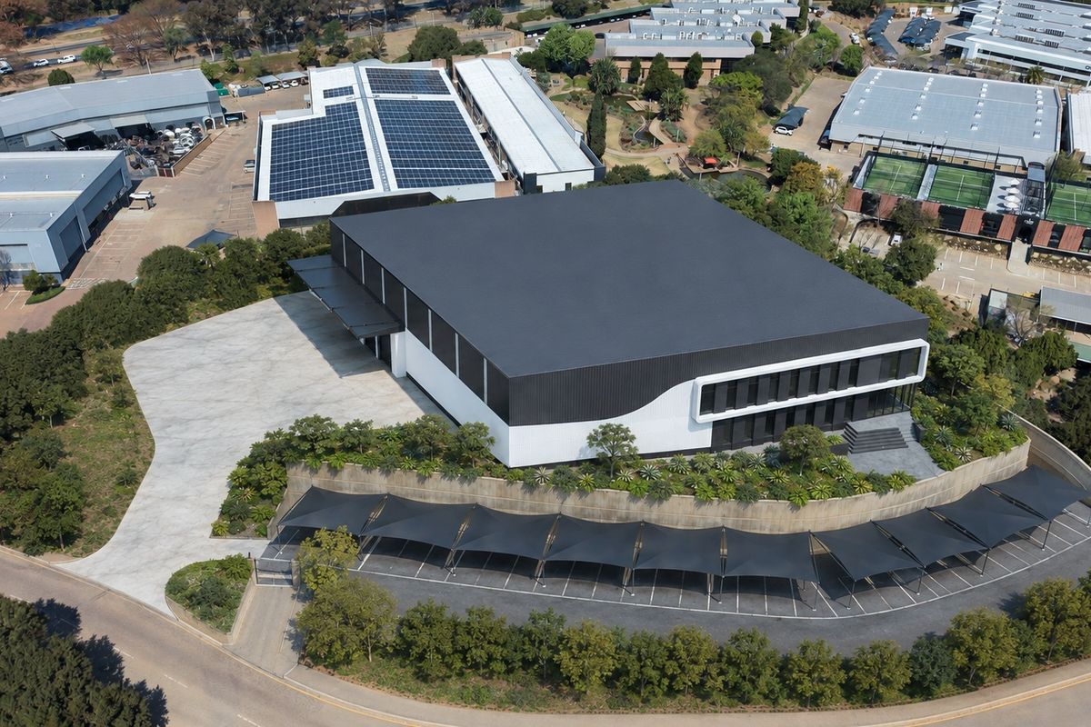 Aerial view of a modern industrial facility featuring a large warehouse with solar panels, extensive paved parking with shade structures, and landscaped grounds.