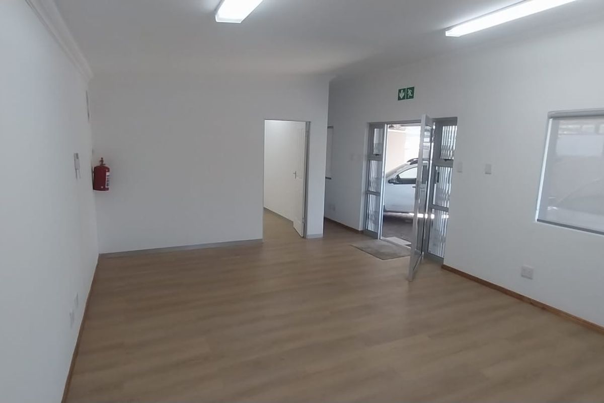A clean, vacant office space featuring laminate flooring, bright fluorescent lighting, and large windows providing natural light and exterior access.