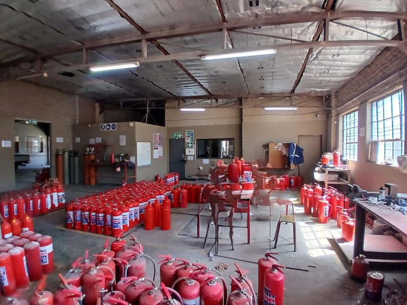 Industrial warehouse or utility space featuring exposed steel roof trusses, concrete flooring, and large steel-framed windows, currently utilized for fire extinguisher storage.