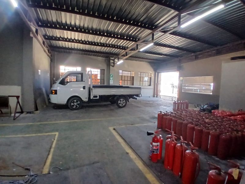 Spacious industrial warehouse or garage area featuring high corrugated metal ceilings, exposed steel trusses, concrete flooring, and ample natural light through industrial windows.
