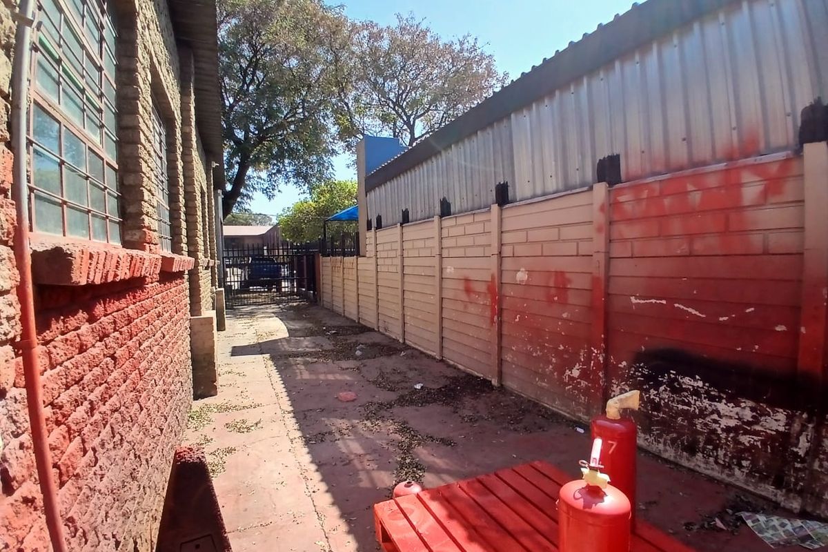 A narrow exterior side passage between industrial buildings featuring brick and corrugated metal walls, a paved walkway, and a perimeter boundary wall.
