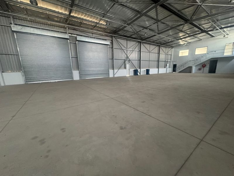Spacious industrial warehouse featuring high ceilings with exposed steel trusses, concrete flooring, and multiple large roller shutter doors for easy access.
