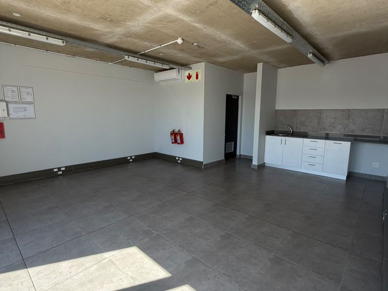 An open-plan industrial office space featuring exposed concrete ceilings, tiled floors, a kitchenette with built-in cabinetry, and integrated air conditioning.