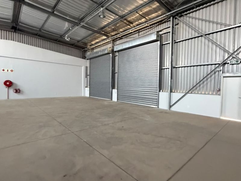 Spacious industrial warehouse unit featuring high ceilings with exposed steel trusses, concrete flooring, and large roller shutter doors for easy access.