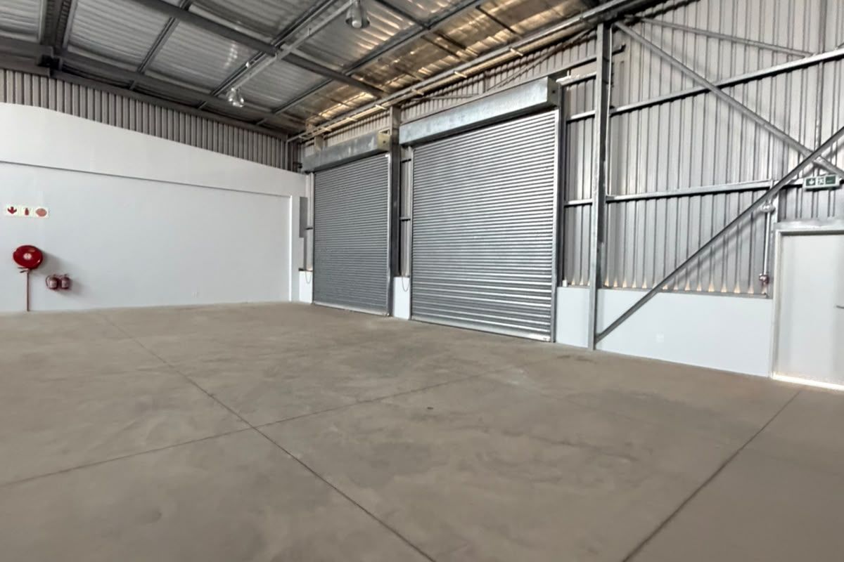 Spacious industrial warehouse unit featuring high ceilings with exposed steel trusses, concrete flooring, and large roller shutter doors for easy access.