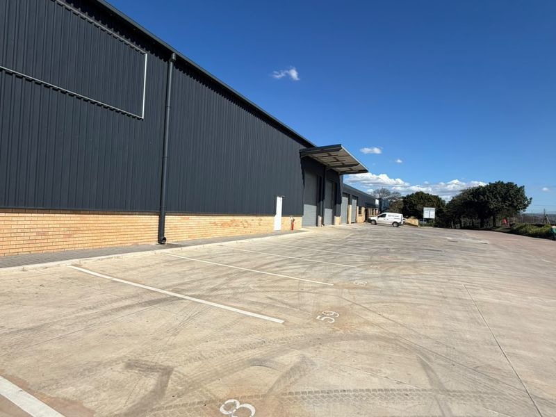 Exterior view of an industrial warehouse facility featuring metal cladding, a brick base, and a paved concrete parking area with marked bays.