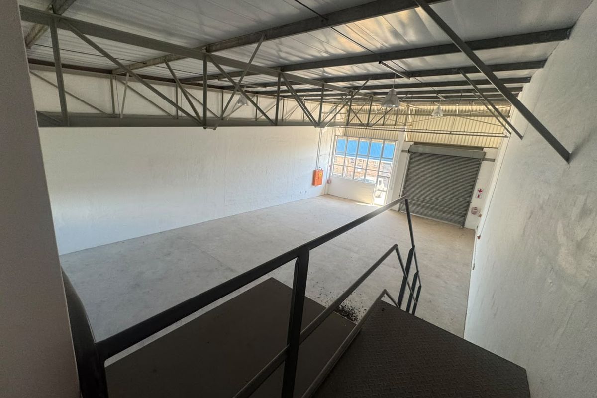 Spacious industrial warehouse unit featuring high exposed steel trusses, concrete flooring, a roller shutter door, and an internal metal staircase.