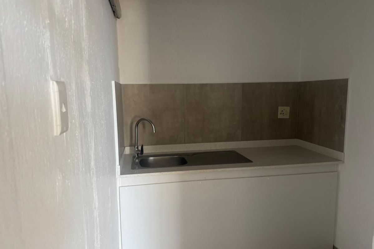 A compact kitchenette area featuring a stainless steel sink, neutral tiled backsplash, and white base cabinetry, suitable for a small utility or office space.