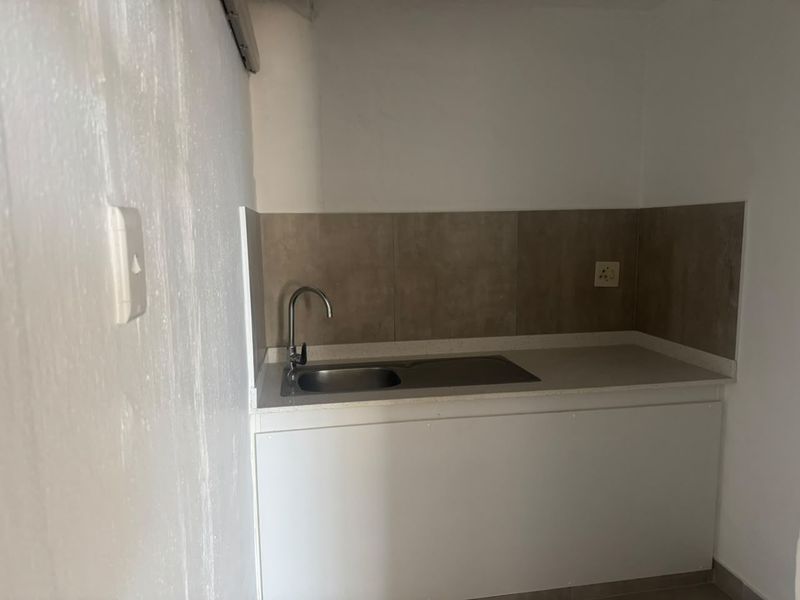 A compact kitchenette area featuring a stainless steel sink, neutral tiled backsplash, and white base cabinetry, suitable for a small utility or office space.