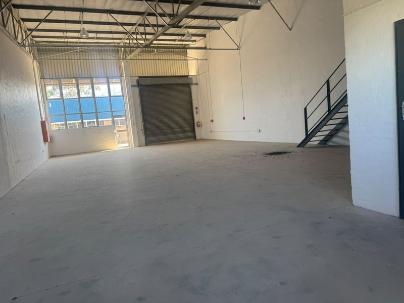 Spacious industrial warehouse unit featuring high ceilings with exposed steel trusses, concrete flooring, a roller shutter door, and an internal metal staircase.