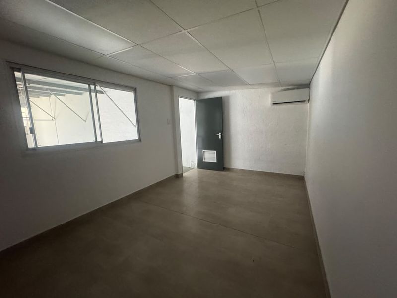 A clean, empty office space featuring a suspended ceiling, tiled flooring, a sliding window, and a wall-mounted split-system air conditioning unit.
