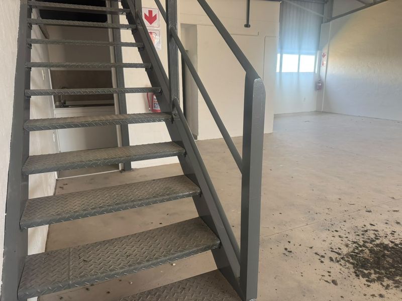 Industrial utility space featuring a metal staircase, concrete flooring, and a small kitchenette area, suitable for various commercial or storage purposes.