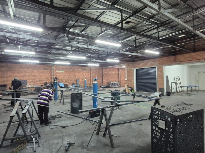 Spacious industrial workshop featuring exposed brick walls, high steel truss ceilings, fluorescent lighting, and a large roller shutter door for easy access.