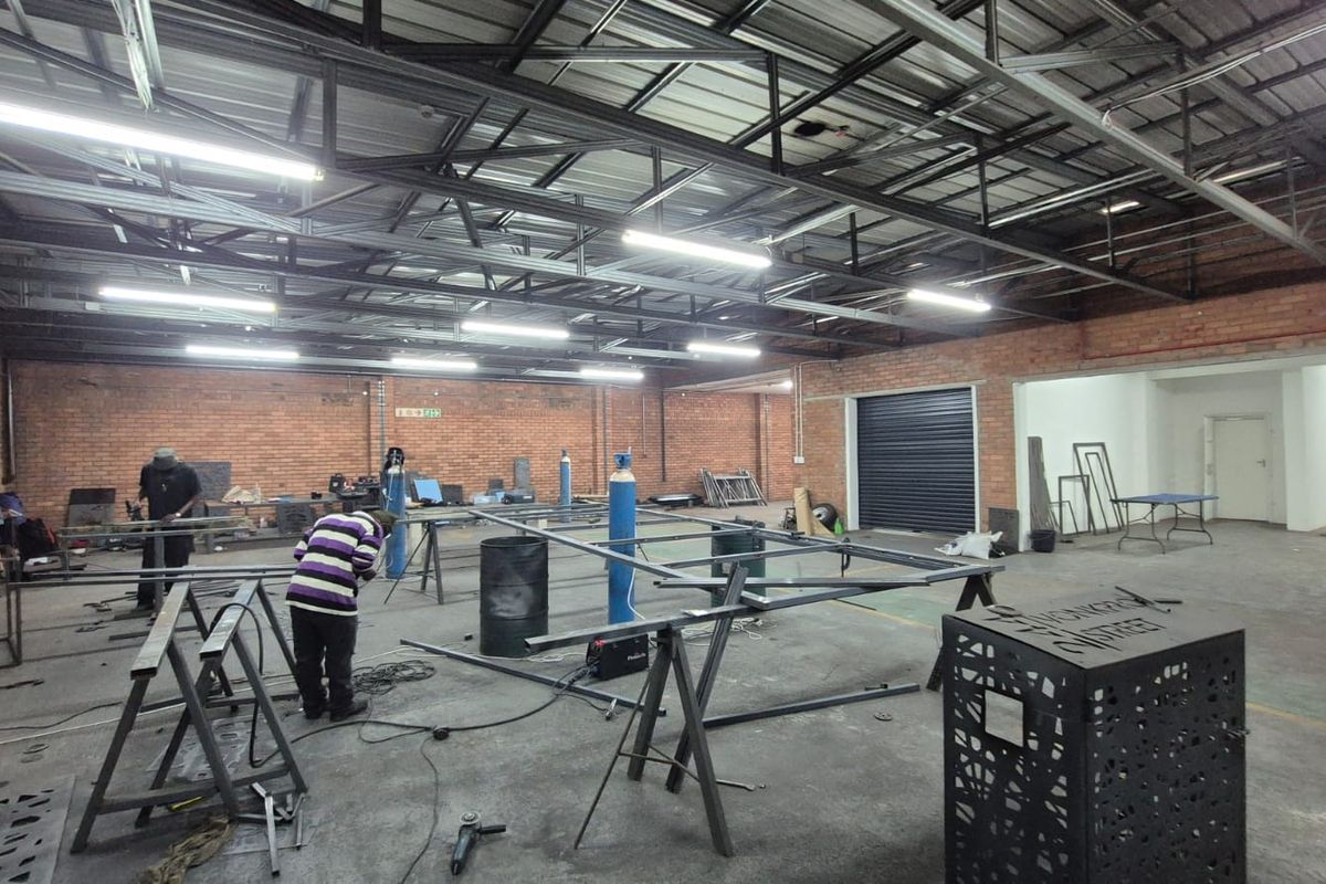 Spacious industrial workshop featuring exposed brick walls, high steel truss ceilings, fluorescent lighting, and a large roller shutter door for easy access.