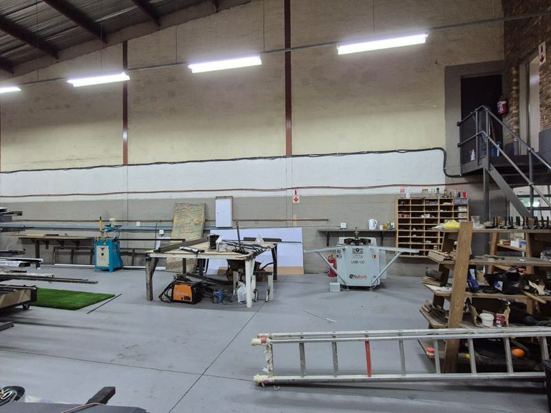 Spacious industrial workshop featuring high ceilings with exposed steel trusses, concrete flooring, and a mezzanine level accessed by a metal staircase.