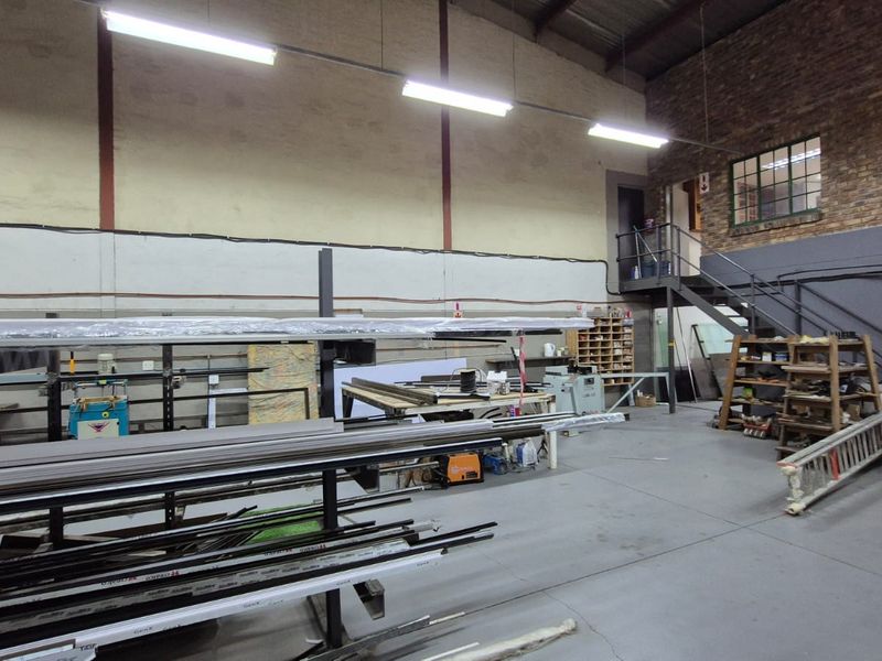 Industrial warehouse space featuring high ceilings with exposed steel trusses, concrete flooring, a mezzanine level, and functional metal staircase access.