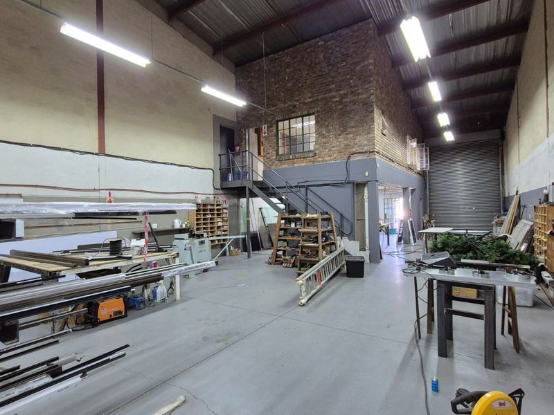 Spacious industrial warehouse unit featuring high ceilings with exposed trusses, concrete flooring, a roller shutter door, and an internal mezzanine level with staircase.