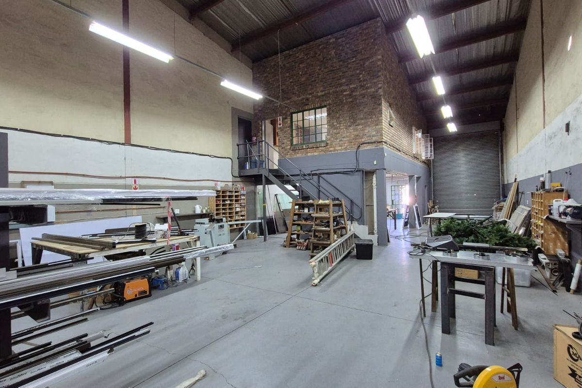 Spacious industrial warehouse unit featuring high ceilings with exposed trusses, concrete flooring, a roller shutter door, and an internal mezzanine level with staircase.