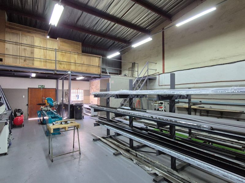 Industrial workshop space featuring a mezzanine level, exposed steel roof trusses, fluorescent lighting, and durable concrete flooring suitable for manufacturing or storage operations.