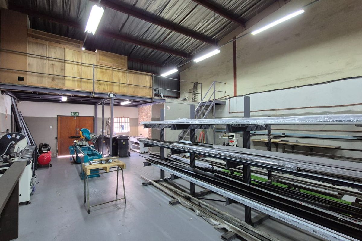 Industrial workshop space featuring a mezzanine level, exposed steel roof trusses, fluorescent lighting, and durable concrete flooring suitable for manufacturing or storage operations.