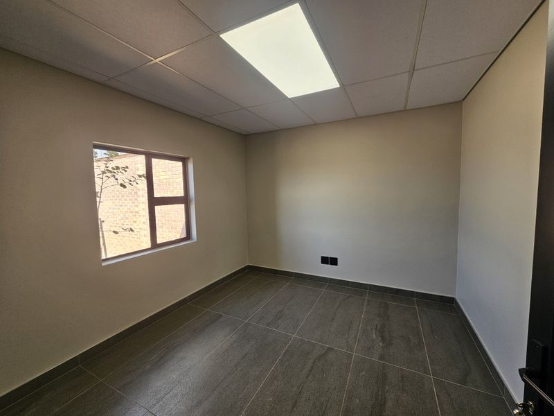 A clean, empty office space featuring modern tiled flooring, a suspended ceiling with recessed lighting, and a window providing natural light.