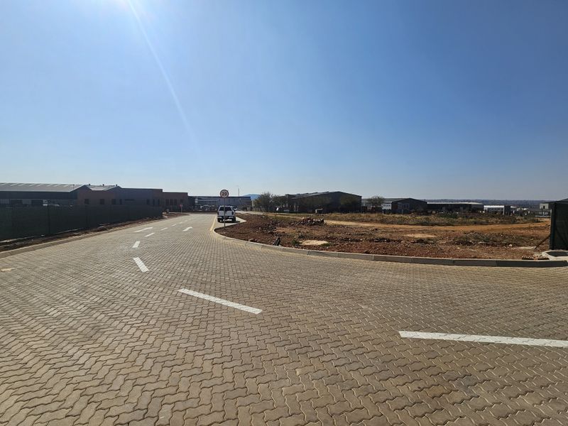 A view of a paved industrial estate road with clear road markings, concrete curbing, and surrounding undeveloped land under a clear blue sky.