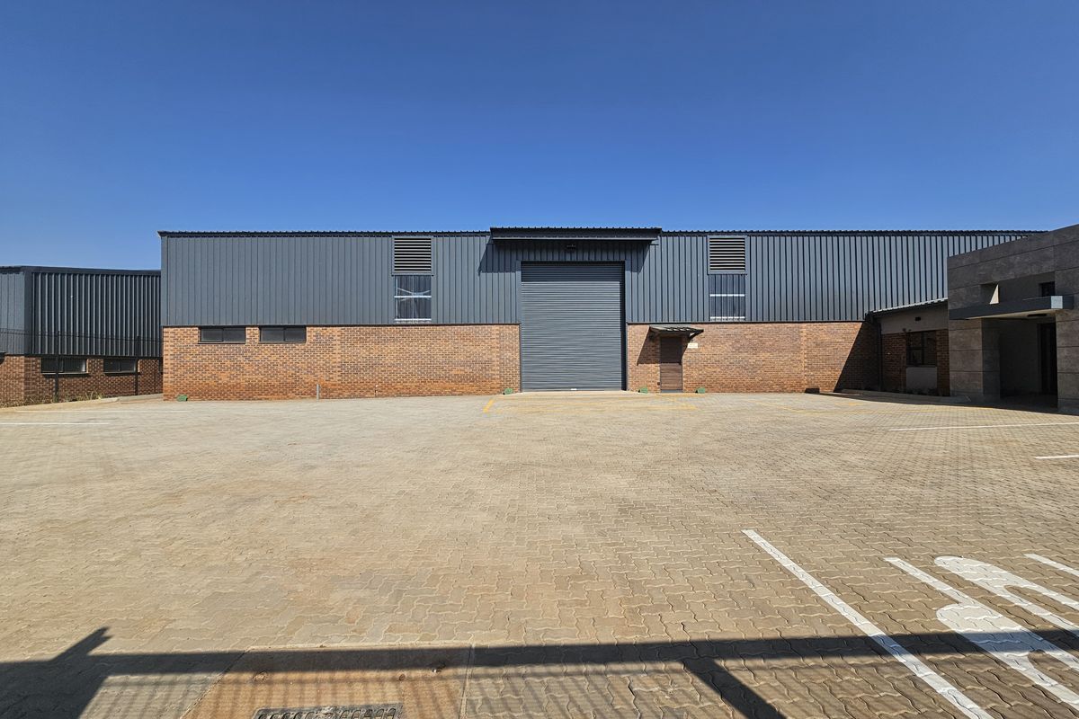 Industrial warehouse exterior featuring a large roller shutter door, brick and metal cladding facade, and a spacious paved parking area.