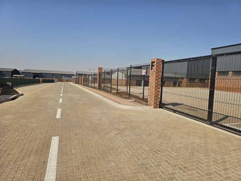 View of an industrial park entrance featuring paved access roads, secure perimeter fencing with brick pillars, and modern warehouse structures.