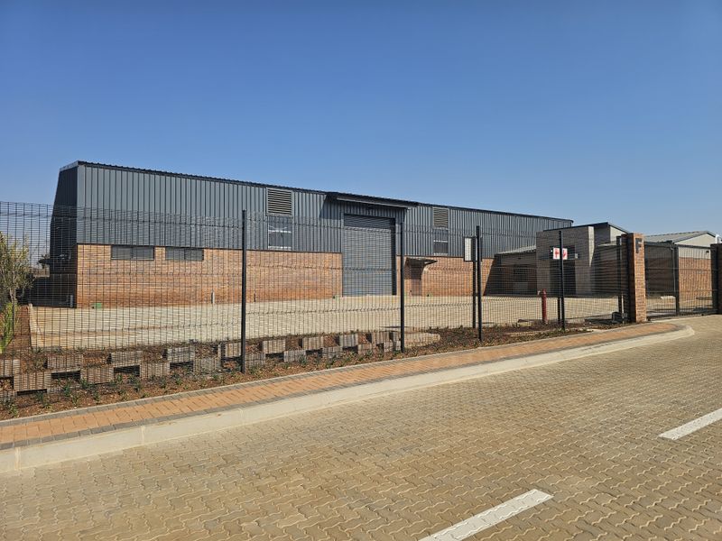 Front view of a modern industrial warehouse unit featuring a face brick base, metal cladding, roller shutter door, and secure perimeter fencing.