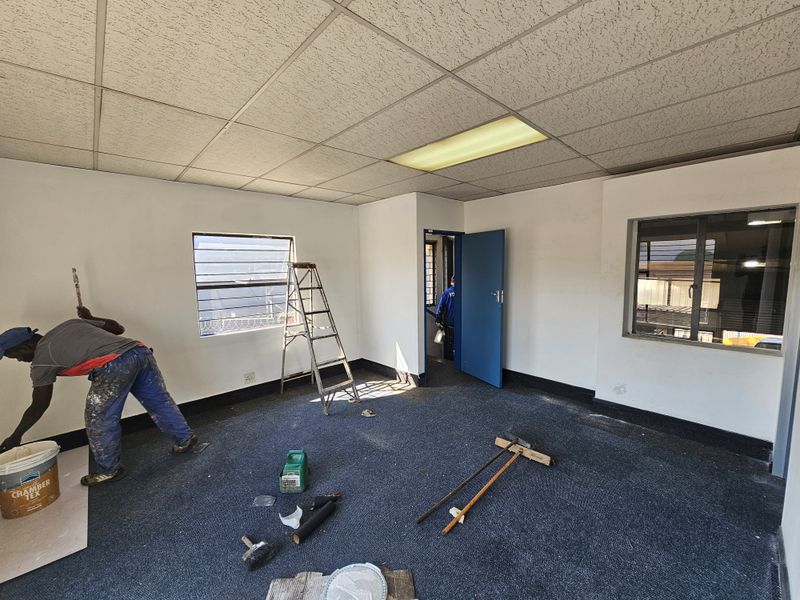 An industrial office space undergoing maintenance, featuring a suspended ceiling, recessed lighting, and louvered windows, currently being repainted.