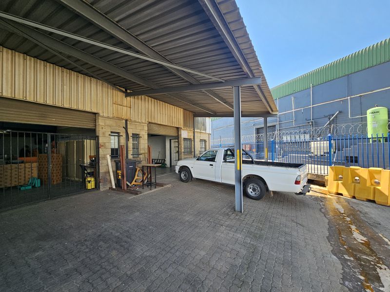 Industrial rear exterior area featuring a covered loading bay with roller shutter doors, paved driveway, and secure perimeter fencing.