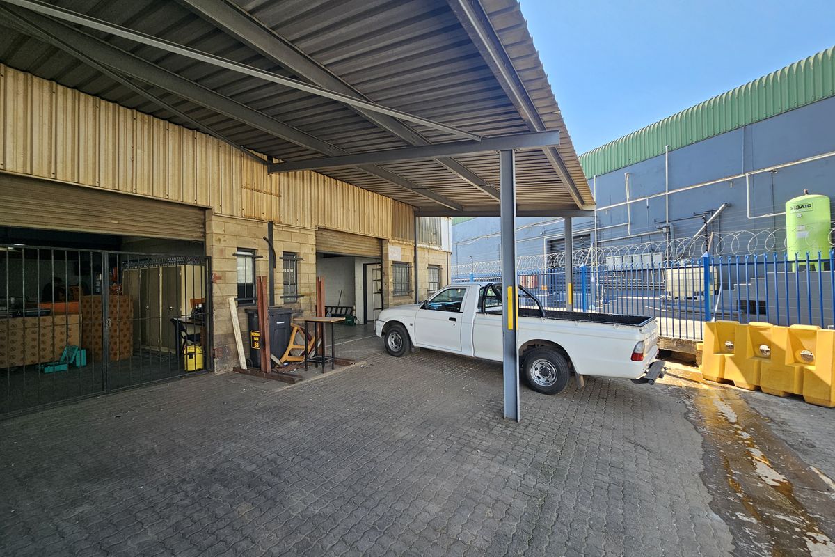 Industrial rear exterior area featuring a covered loading bay with roller shutter doors, paved driveway, and secure perimeter fencing.