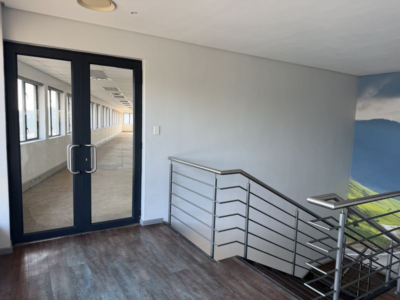 A bright office hallway featuring a modern stainless steel balustrade, laminate flooring, and double glass doors leading into an open-plan workspace.