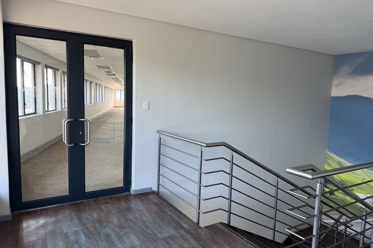 A bright office hallway featuring a modern stainless steel balustrade, laminate flooring, and double glass doors leading into an open-plan workspace.