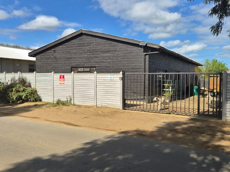 Front exterior view of an industrial property featuring a dark-painted concrete block building, secure metal gate, and precast concrete perimeter fencing.